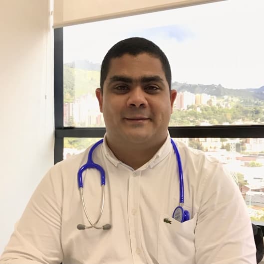 Smiling male doctor in a white shirt with a blue stethoscope and city background.