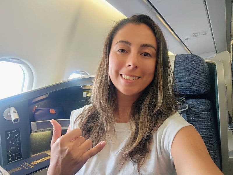 Smiling woman sitting in an airplane business class seat making a shaka hand gesture.