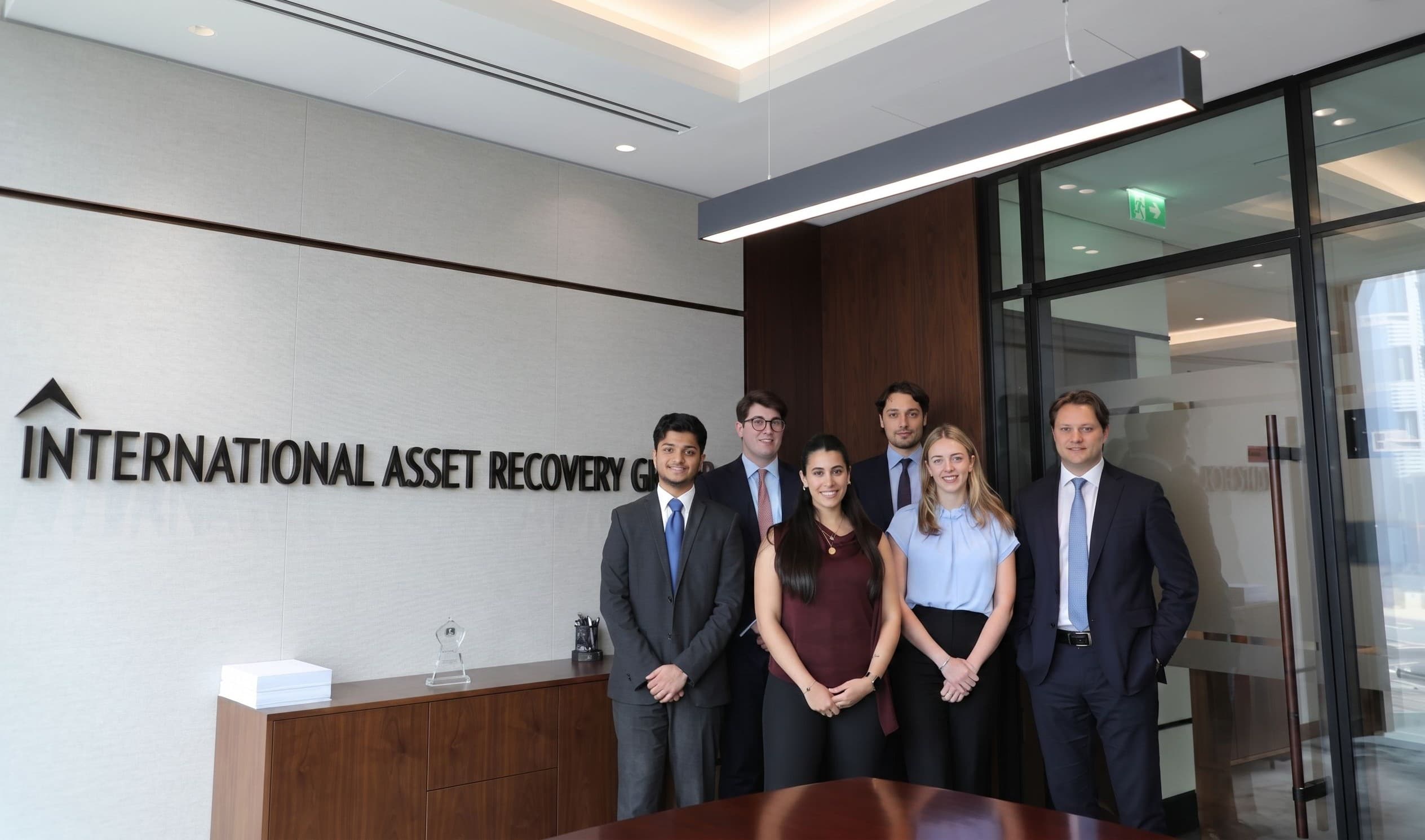 Six professionals in business attire posing before the International Asset Recovery Group office sign.